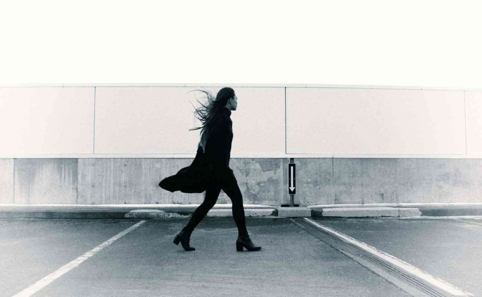 Death and life with chronic illness. A woman in boots and a long black coat walks across a parking lot.