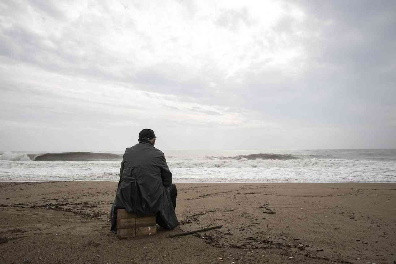 Loneliness and schizophrenia. A sad man sitting alone on the beach with stormy skies above.