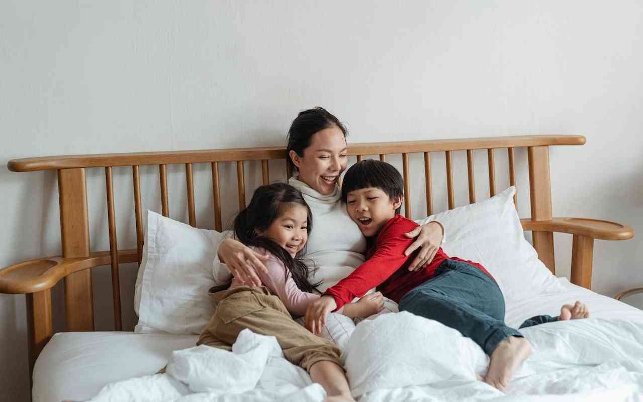 Parenting with chronic fatigue. A mother of Asian heritage hugs her kids while resting in bed.