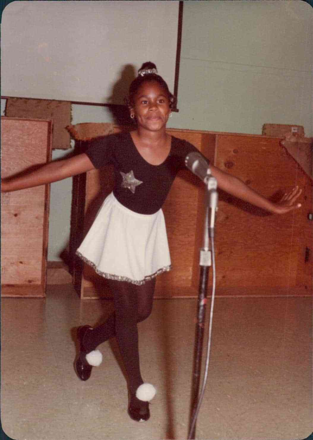 Alisa as a child, wearing a ballet outfit and standing in front of a microphone.