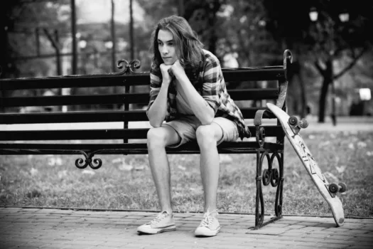 Sad teen with skateboard sitting on a park bench.