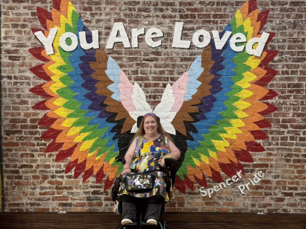 Karin Willison. A white woman with long blonde hair sits in a power wheelchair. She is wearing a rainbow dress with rainbow angel wings painted on the brick wall behind her.